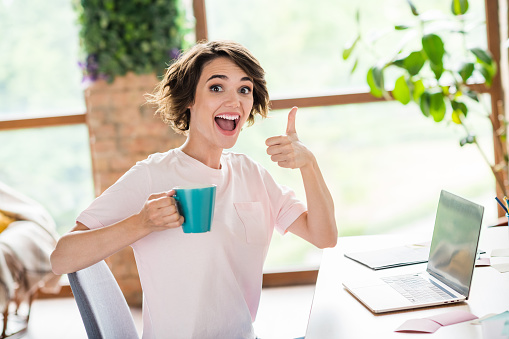 Photo of excited surprised young beginner intern girl day in office thumb up rate her workplace drink tea while studying laptop indoors.