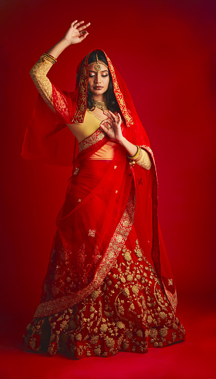Fashion. dance and Indian woman in a traditional dress, jewellery and celebration on a red studio background. Female person, girl and model with a cultural outfit, dancing and routine with confidence