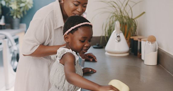 Little girl helping her mother with household chores at home. Happy mom and daughter wearing gloves while spraying and scrubbing the kitchen counter together. Kid learning to be responsible by doing tasks