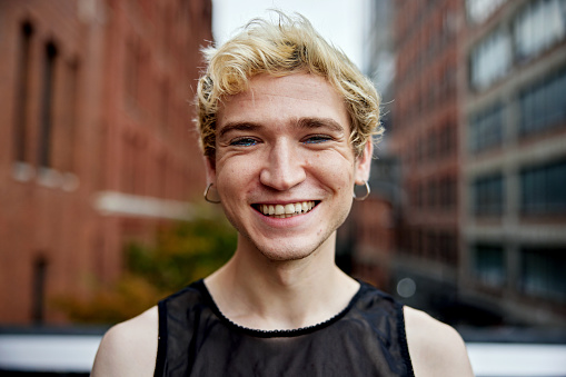 Close front view of gay man with short bleached blond hair, wearing black tank top and earrings, standing outdoors in Chelsea.
