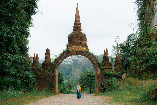 Female traveler in straw hat walking near the gate that leads to an ancient temple in the jungles of Thailand