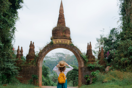 Female traveler in straw hat walking near the gate that leads to an ancient temple in the jungles of Thailand