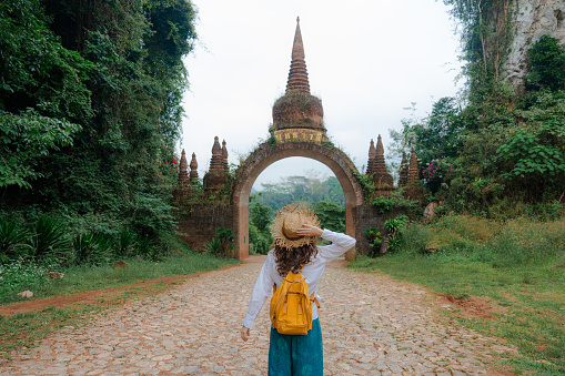 Female traveler in straw hat walking near the gate that leads to an ancient temple in the jungles of Thailand