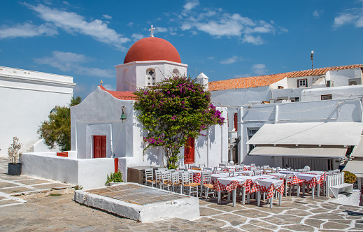 Travel image with white church and red dome in the city center of Mykonos near square with outdoor restaurant tables and chairs without people on a beautiful summer day.