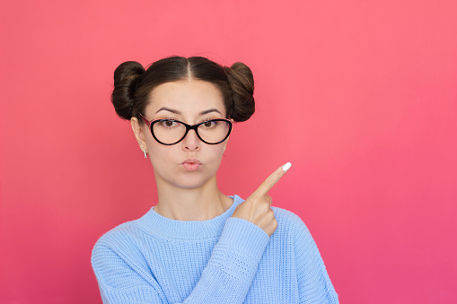 a young pretty brunette woman with glasses, hair and blue clothes with a serious face points with her index finger at an empty background. Isolated on a pink background. Copy space