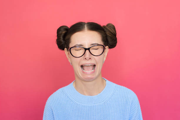 une jeune jolie femme vêtue de bleu, portant des lunettes, est très bouleversée, pleure et crie la bouche ouverte. - mouth open women displeased anger photos et images de collection