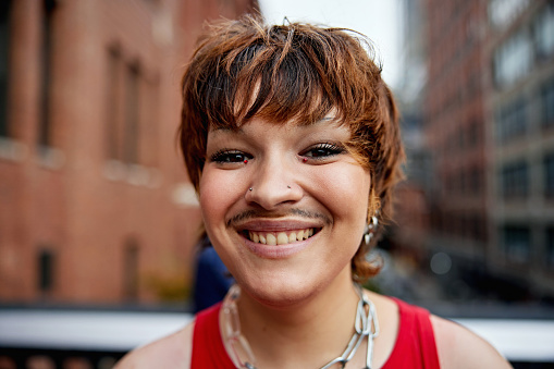 Headshot of happy individual with tousled brown hair, mustache, wearing red tank top, jewelry, and grinning at camera with Chelsea architecture in background.