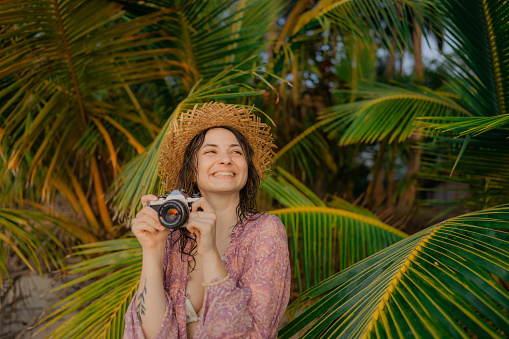 Woman in straw hat making memories photographing on vintage camera during beach vacation in tropical climate