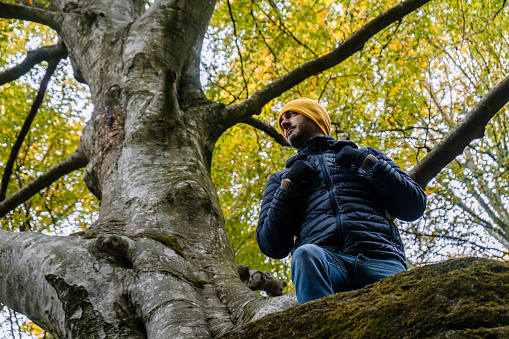 View from below of a handsome man dressed with winter outerwear clothes, looking away standing in the forest in front of a big tree. Hiking in a beautiful landscape during autumn, winter.