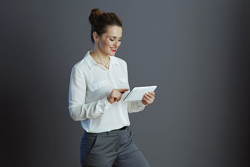 happy trendy middle aged business woman in white blouse using applications on tablet PC against gray background.