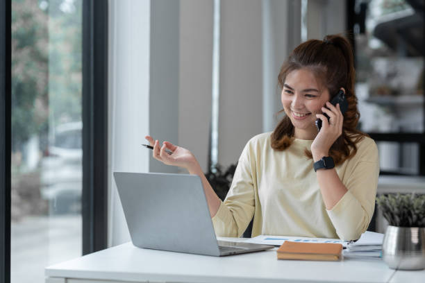 Asian businesswoman or freelancer using laptop computer and mobile phone to communicate with colleague in explaining online meeting or online training. office paperwork
