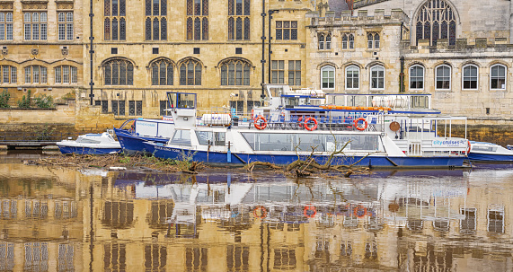 York, UK. October 24, 2023.  Historic buildings and a rows of boats are reflected in a river.  Tree debris from a recent storm float near a tour boat.