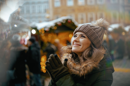 smiling young woman in green coat and brown hat at the winter fair in the city warming hands.