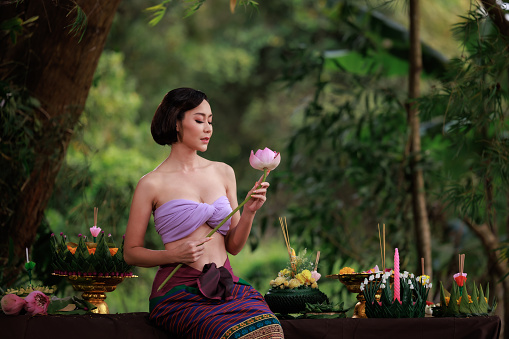 Beautiful young woman in traditional Thai  dress sitting and holding lotus flower in her hands to decorate krathong in the nature background, in loy krathong day,