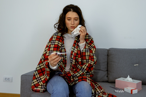 Desperate young Caucasian ill woman holding a paper tissue in one, and thermometer in other hand while sitting on sofa