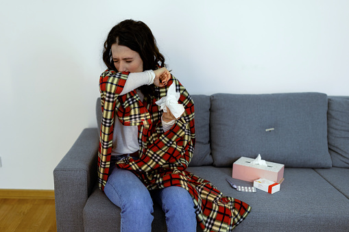Ill young Caucasian woman struggling with cold and flu, sitting on a living room couch covered in blanket and blowing her nose