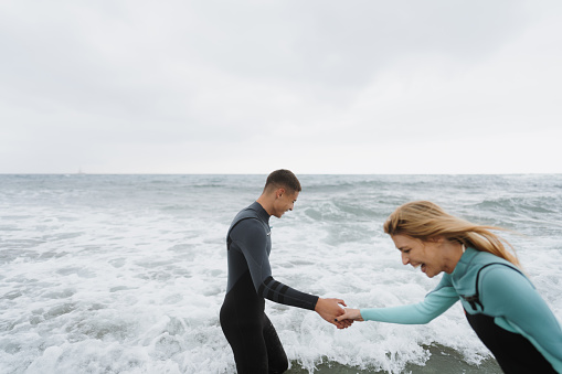 Photo of a couple of smiling people in wetsuits having fun while trying to get into a cold ocean water