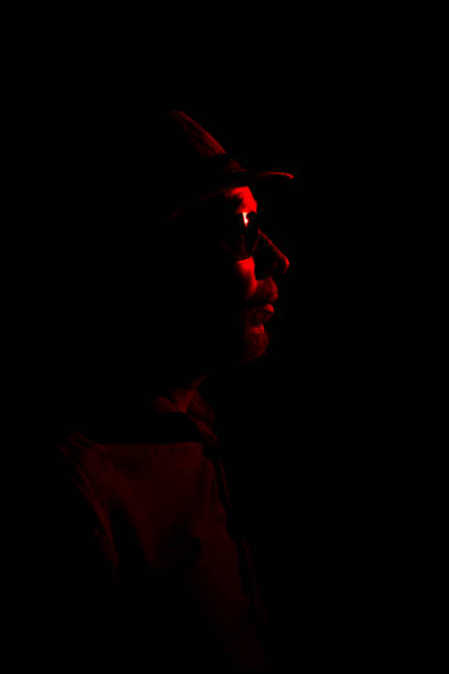 istock Man with hat on dark background, wearing sunglasses with red light on his face. 1779918474