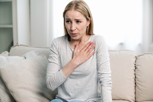worried woman feeling acute cardiac pain, hold hand on chest and having difficulty breathing while sitting on couch at home. young female have panic attack and afraid for her health