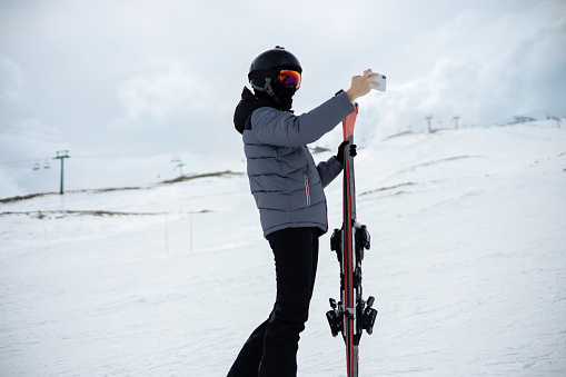 Girl dressed in a ski suit and helmet with balaclava stands with ski against the backdrop of snow-covered mountain ski slope and a cloudy sky and take a selfie on a smartphone.