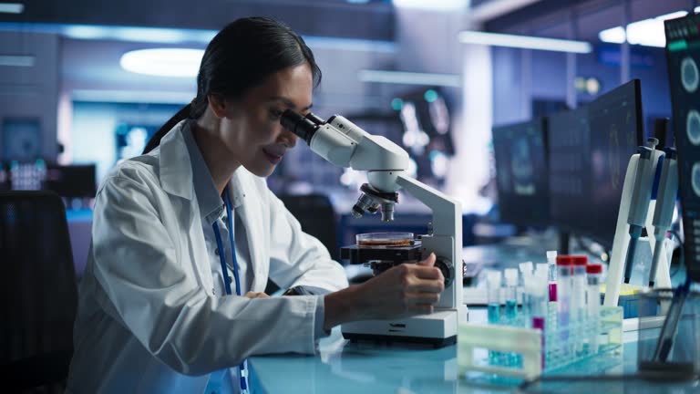 Medical Development Laboratory: Asian Female Scientist Using Microscope, Analyzes Petri Dish Sample. Big Pharmaceutical Lab with Specialists Conducting Biotechnology Research, Developing New Drugs.