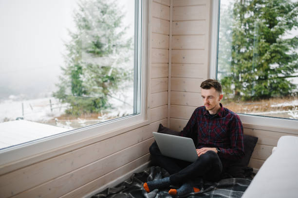 istock Man working on laptop in front of panoramic window with great view of mountains. Workplace in house in snowy morning. Remote work from cozy place. Wide interior view. Concept freelance lifestyle. 1780022963