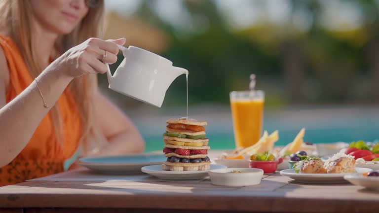 Woman pouring honey on pancakes, pancakes, Sweet honey is poured on pancakes, Young woman having breakfast by the pool at a hotel facility on a sunny summer day, summer breakfast, enjoying a poolside breakfast-dining outside, rich breakfast menu