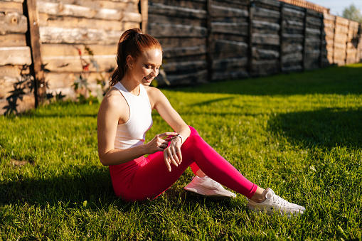 Portrait of happy female in sportwear smiling looking smart watch screen sitting on lawn with green grass on sunny summer day. Redhead young sportswoman using checking fitness tracker outdoors.
