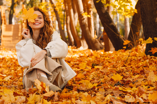 Smiling young woman sits in the autumn foliage on the ground of the forest with the yellow leaves