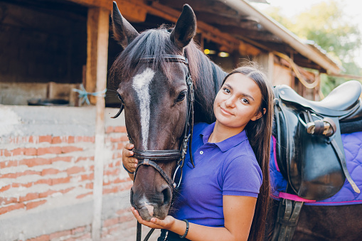 Young woman with her horse in a barn. She is petting him and smiling.
