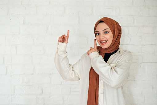 Woman wearing hijab pointing hands upwards and smiling at camera, standing against white brick wall