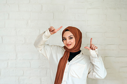 Woman wearing hijab looking at camera, smiling and pointing hands upwards, standing against white brick wall