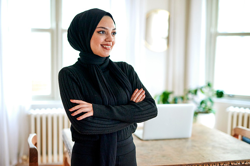 Young woman with hijab wearing black smiling into distance and standing with arms crossed in home office