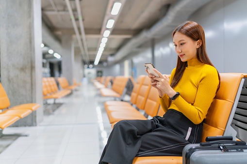 Smiling young Asian woman reading on a mobile phone while waiting for her flight in airport lounge at night. Asian businesswoman working on a smartphone. Asian woman on business trip. Lifestyle and technology. Modern travel and vacation concept. Business on the go.