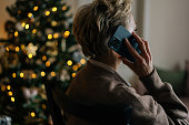 Rear view of unrecognizable woman chatting smartphone in dark room by illuminated Christmas tree. Happy female enjoying pleasant phone call,