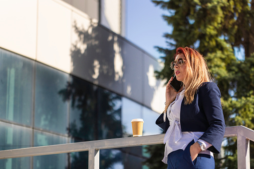 Modern mid-adult Caucasian businesswoman, drinking coffee and taking on a mobile phone during a coffee break, in front of the corporate building