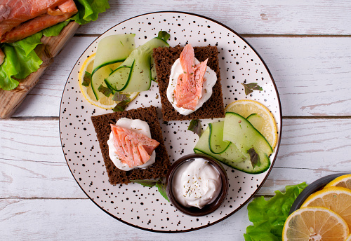 Canape with hot smoked red fish and sour cream sauce on rye bread served with cucumber and lemon slices on white plate. Top view