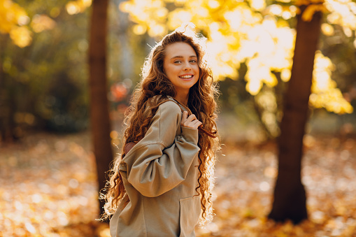 Smiling young woman walking in autumn forest with the yellow leaves at sunset