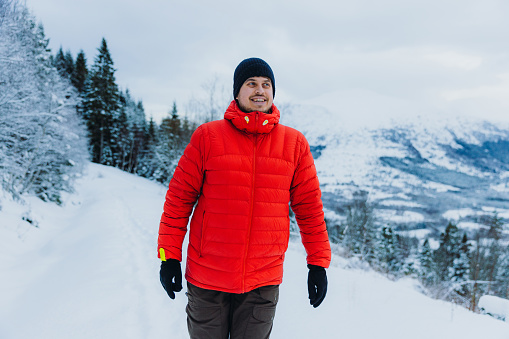 Front View of a smiling man in red jacket contemplating the Christmas time outdoors walking in the deep snow in pine woodland with Mountain View of Norway, Scandinavia