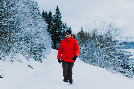 Front View of a smiling man in red jacket contemplating the Christmas time outdoors walking in the deep snow in pine woodland with Mountain View of Norway, Scandinavia