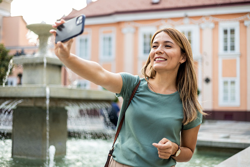 Young Caucasian woman taking a selfie in town in a busy street posing for the camera on her smartphone with a smile.
