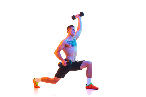 Athletic young man with strong, relief shirtless body doing lunges with dumbbells against white studio background in neon light. Concept of sport, active and healthy lifestyle, body care, fitness