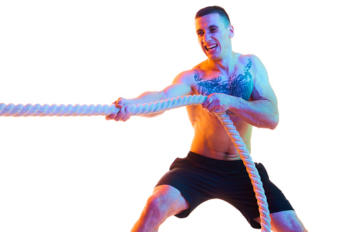 Shirtless athletic young man with strong body and hands pulling rope, training against white studio background in neon light. Concept of sport, active and healthy lifestyle, body care, fitness