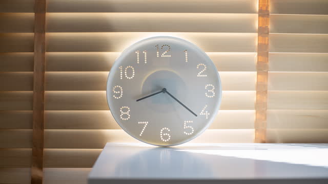 Sunlight shining through window blinds on a white clock sitting on the table inside an office and the light is getting brighter as the time passes by during the day.