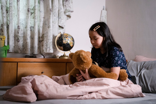The Asian girl cuddling her bear doll sitting alone on the bed in the room