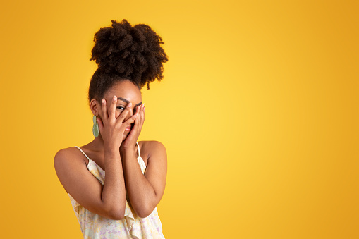 Smiling embarrassed shy millennial african american lady in dress covering face with hands, isolated on yellow background, studio. Surprise, fashion lifestyle, beauty care, holiday, ad and offer