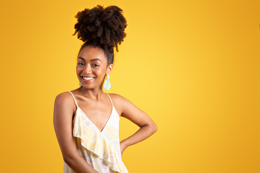 Cheerful confident young black woman in dress looks at camera, enjoys vacation, weekend and spare time, isolated on yellow background, studio. Lifestyle, fashion sale, ad and offer
