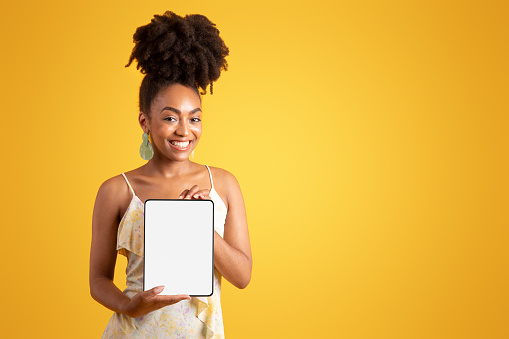 Cheerful young black woman showing tablet with empty screen, recommending social media app, isolated on yellow background, studio. Website, ad and offer, lifestyle, blog and device for work, study