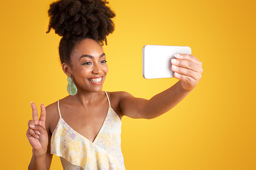 Cheerful young african american lady in dress taking selfie on smartphone, make peace sign, isolated on yellow studio background. Lifestyle, fashion blog, ad and offer, gadget for photo