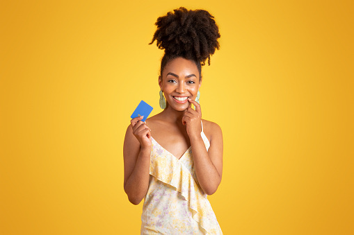 Smiling millennial african american lady showing credit card, recommends finance savings, isolated on yellow background, studio. Shopping, ad and offer, money control, pay, buy